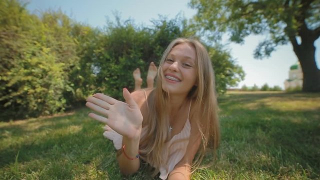 slow motion happy blond long haired girl with dental braces lies on grass makes hand figures in front of camera in park