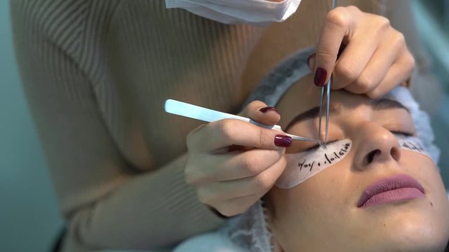Woman is in cosmetologic salon.She is lying with a closed eyes and in hair mask. A beautician is doing the procedure of eyelash extension. With special equipment she is styling lashes.