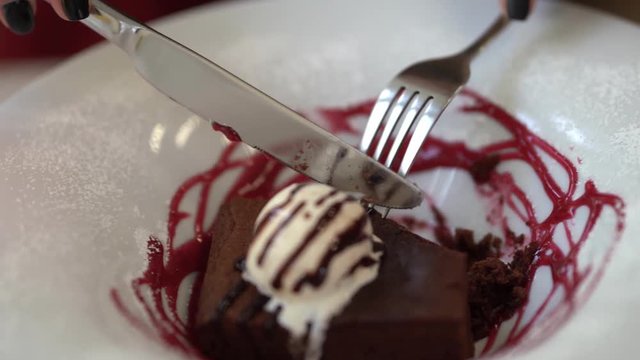 Close scene as a girl is cutting her dessert with fork and knife. There is an ice cream on the top of cake and plate is decoretade with red topping.