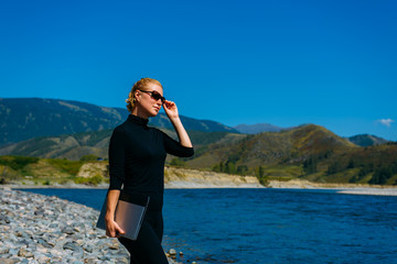 Freelancer with laptop on the nature. Blogger hipster traveler business lady pretty girl   finished work outdoor. Young blondy woman with sunglasses in black standing next to mountain river