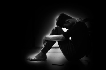 Boy with lower head and arm around knee sitting on corner of room in the dark
