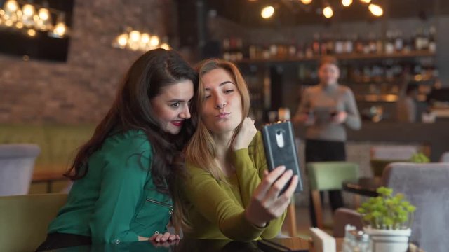 Two beautiful women friends sitting in a cafe, taking photos of themselve, posing for photo, trying to make nice shot, kissing on the cheeck