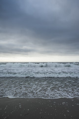Sunset on horizon, ocean waves and sand coastline in front, gray cloudy sky