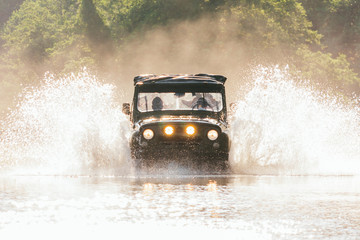 Old classic 4x4 car crossing water with beautiful splashes in a warm sunny day, low angle front view. Extreme off-road tour in a forest