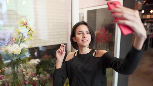 Beautiful slim thin hot sexy brunette in black shoulderless sweater standing near flower shop and filming herself talking on camera, doing story for instagram, pointing to shop window/shopcase