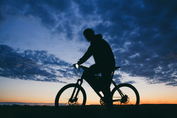 Fototapeta na wymiar Silhouette of a young man in a trip on the mountain bike with head lamp in a dark conditions against a cloudy evening sky . Low angle view with motion blur