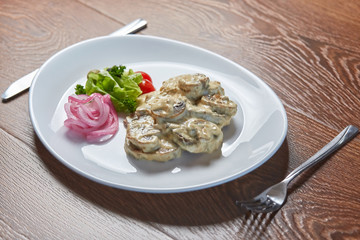 champignons baked in cheese, next is a cut cherry tomato, olives, pickled onions, cilantro and a leaf of salad, all on a white plate standing on a wooden table