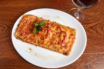 lasagna with tomatoes and minced puff pastry, decorated with slices of cherry tomatoes, parsley leaf and olive, standing next to a glass of red wine