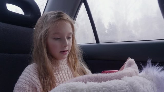 Cute young girl with long hair drive travel in car and use smart phone. White girl in pink warm sweater looking cell phone gadget. White fur jacket winter travel