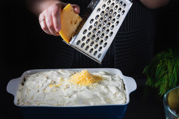 Female chef hands rub cheese on an old grater into baking dish. Ingredients for cooking of potato...