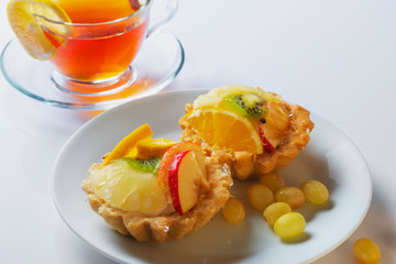 Basket-cake decorated with different fruit and cup of tea on white table.