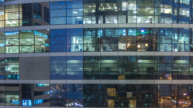 Glowing windows in multistory modern glass and metal office building light up at night timelapse.
