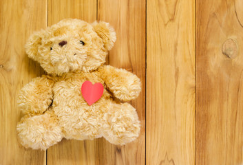 Teddy bear holding red paper heart shape on wooden background.