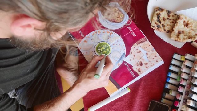 Man sits in cafe and drawing image of Indian food in sketchbook by art markers
