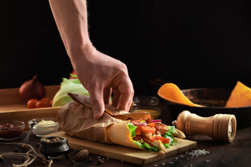 Shawarma sandwich in men hand on wooden rustic background. Gyro fresh roll of lavash or pita bread with grilled chicke, lettuce salad, cheese and vegetables. Tasty shawarma - middle eastern snack.