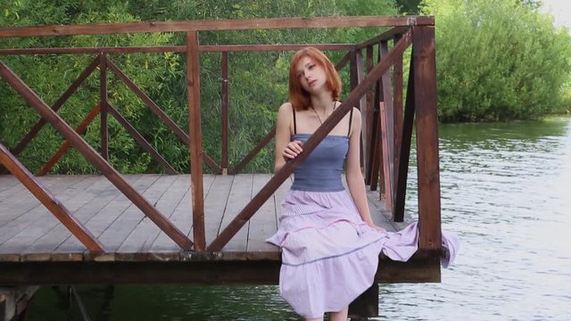 Attractive young woman sitting on pier in summer. Red-haired bright beautiful young woman in long skirt sitting on pier on background of green vegetation
