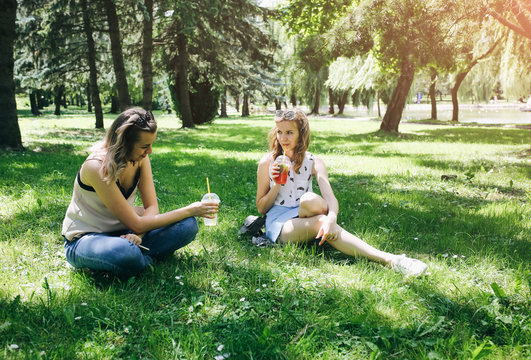 Two hipster girls are laughing and drinking summer cocktails outdoors in the green grass. Cold non-alcoholic drinks with ice to go. Mojito and strawberry lemonade. Happy lifestyle for vacations.