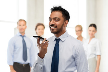 business, communication and technology concept - smiling indian businessman using voice command recorder on smartphone over colleagues on office background