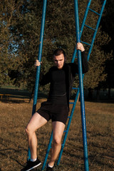 A young muscular man pulls himself up on a bar in the background of the sun on the street.