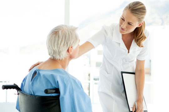 Beautiful Nurse Caring Senior Male Patient Sitting On Wheelchair At The Hospital - Hand On Shoulder - Looking At Each Other