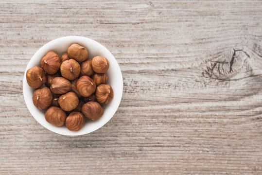 Hazelnuts peeled in white little bowl on wooden background, top view.