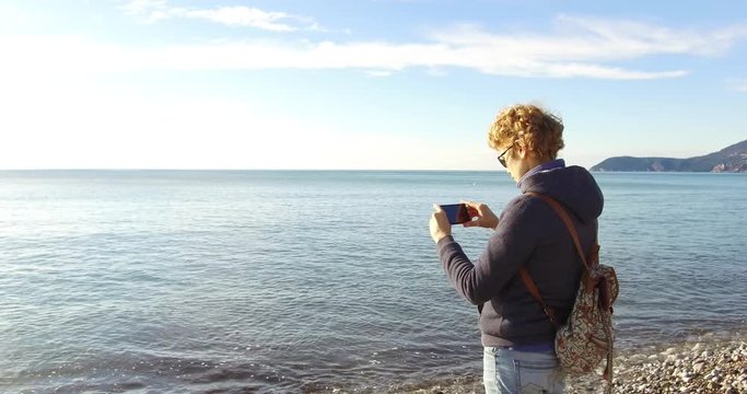 Sunny spring day before sunset. Tourist girl in a warm sweater with a backpack on her back is standing on the pebble beach with a smartphone in her hand and taking pictures of the sea on a smartphone