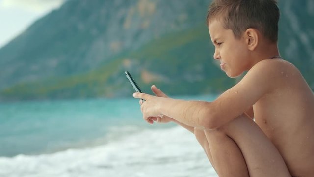 Boy using mobile phone sitting at sea beach. Sea wave hit boy with smartphone
