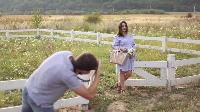 Photographer make a photo for beautiful pregnant woman dressed in the blue dress and knitted hat. She put hands on her belly. Photosession in the field near the farm. Summer time