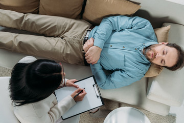 overhead view of psychotherapist writing on clipboard and male patient with closed eyes lying on couch
