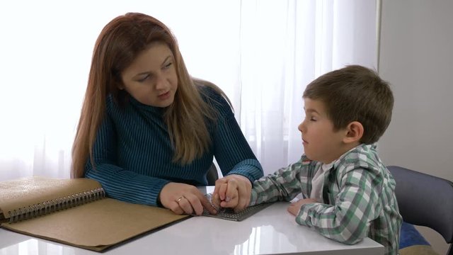 education of blind children, mother teaches child boy to write braille sitting at table in bright room at home