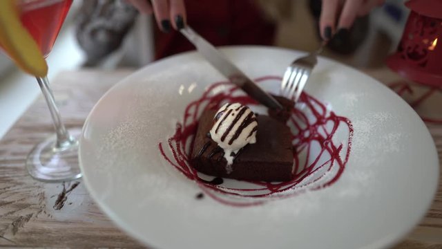 Close scene as a girl is cutting her dessert with fork and knife. There is an ice cream on the top of cake and plate is decoretade with red topping. Also there is a coctail on the table.