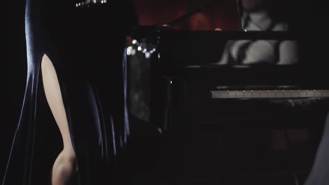 A girl in a beautiful evening dress with a glass of wine comes to the piano where the pianist sits