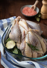 Raw guinea fowl in transparent glass baking dish with garlic and rosemary and next to it are Himalayan pink salt in a bowl and a pepper mill