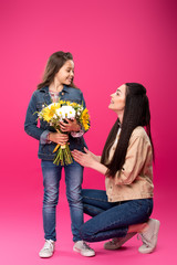 full length view of happy mother looking at cute smiling daughter holding flower bouquet on pink