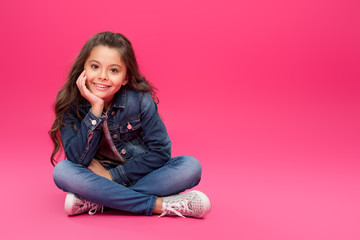 adorable happy child in denim clothes sitting with hand on chin and smiling at camera on pink