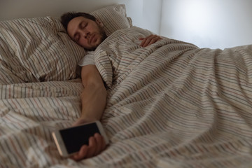 Portrait of dressed adult male fell asleep in bed with the phone in hand as a result of tiredness/ selective focus on man/ social media addiction, accumulated fatigue, not enough sleep.