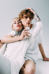 man with curly hair holding cigarette in mouth near blonde tattooed girl isolated on grey