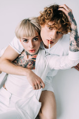 handsome man with curly hair holding cigarette in mouth while sitting with blonde tattooed girl isolated on grey