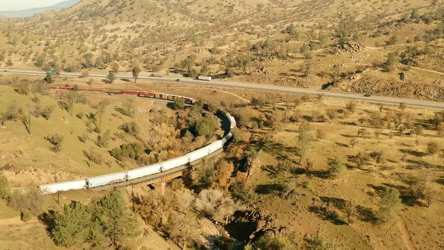 The aerial camera dives in toward a long freight train passing through the northeastern exit of the railroad loop site. This clip shows cars, trucks and Semi-Haulers driving along Highway 85.