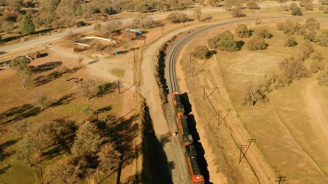 The aerial camera flies 155 feet above three locomotives coming out from the western side of the rail loop tunnel. Camera follows the locomotives curving the tracks to the east along highway 58.