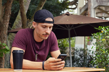 Portrait of serious busy European entrepreneur in t-shirt and hat, using mobile phone for business correspondence, looking at the screen with concentrated look. Face expressions, emotions concept.