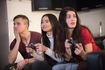 Company of young friends spending time together. Two young woman playing game using a joystick and young man gasp