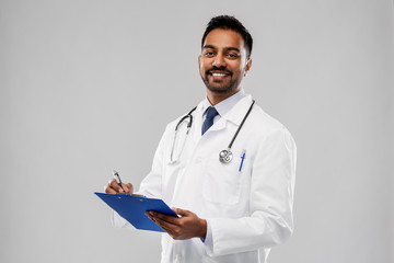 medicine, profession and healthcare concept - smiling indian male doctor in white coat with stethoscope and clipboard over grey background