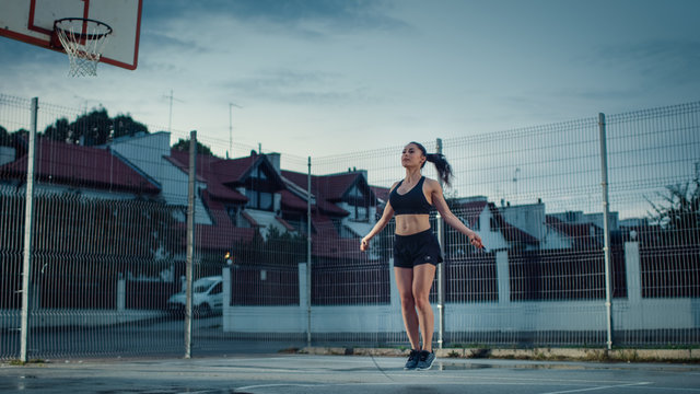 Beautiful Energetic Fitness Girl Skipping/Jumping Rope. She is Doing a Workout in a Fenced Outdoor Basketball Court. Evening Shot After Rain in a Residential Neighborhood Area.