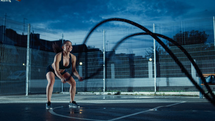 Beautiful Energetic Fitness Girl Doing Exercises with Battle Ropes. She is Doing a Workout in a Fenced Outdoor Basketball Court. Evening After Rain in a Residential Neighborhood Area. © Gorodenkoff