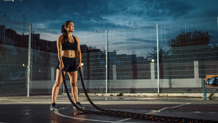 Obraz na płótnie Canvas Beautiful Energetic Fitness Girl Doing Exercises with Battle Ropes. She is Doing a Workout in a Fenced Outdoor Basketball Court. Evening After Rain in a Residential Neighborhood Area.