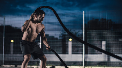 Strong Muscular Fit Shirtless Young Man is Doing Exercises with Battle Ropes. He is Doing a Workout in a Fenced Outdoor Basketball Court. Evening After Rain in a Residential Neighborhood Area.