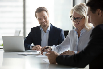 Smiling middle aged older businesswoman signing paper contract at group meeting, happy mature...