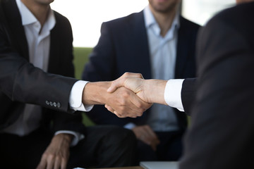 Close up view of two businessmen in suits shake hands at group meeting, business partners handshaking after successful negotiation closing good deal, respect and loyalty, union, reliability concept