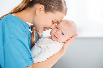 side view of happy young mother carrying and hugging adorable baby looking at camera in hospital room